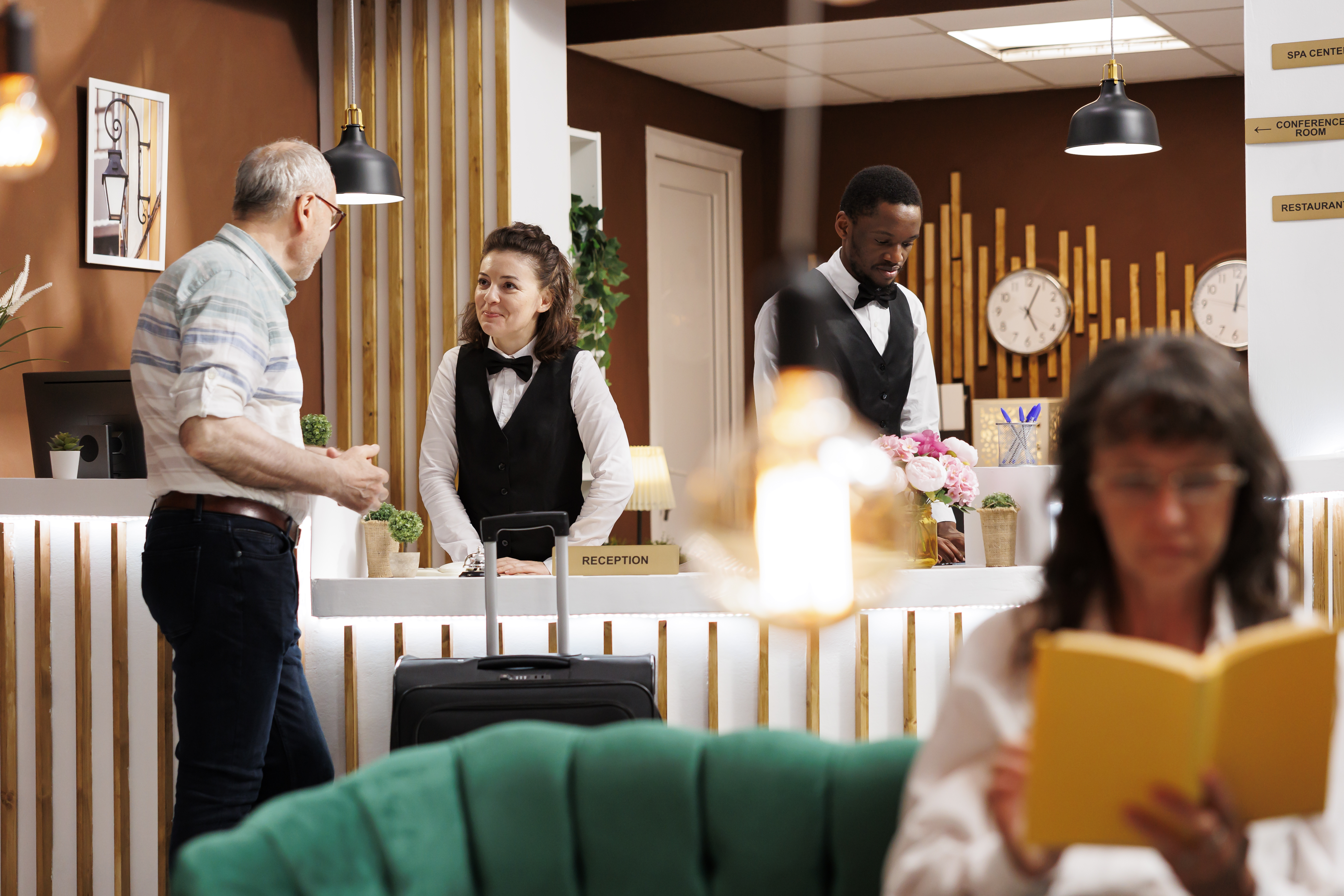 Smiling Concierge Assists Elderly Male Guest Front Desk While Caucasian Senior Woman Enjoys Reading Novel Lounge Area Hotel Lobby Features Comfy Sofa Reception Counter Reservations