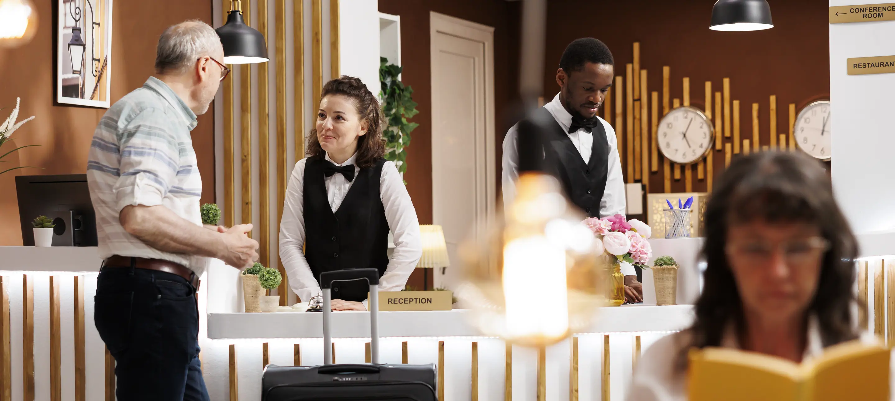 Smiling Concierge Assists Elderly Male Guest Front Desk While Caucasian Senior Woman Enjoys Reading Novel Lounge Area Hotel Lobby Features Comfy Sofa Reception Counter Reservations